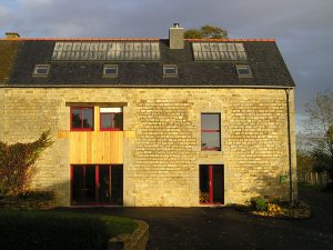 photo d'une maison en pierre avec une portes vitrée , une baie vitrée et deux fenêtres côte à côte le tout cintrées de rouge, quatre lucarnes de toit, avancée goudronnée, mur de délimitation en pierres, du gîte de groupe et maison d'hôtes du Bas Mena