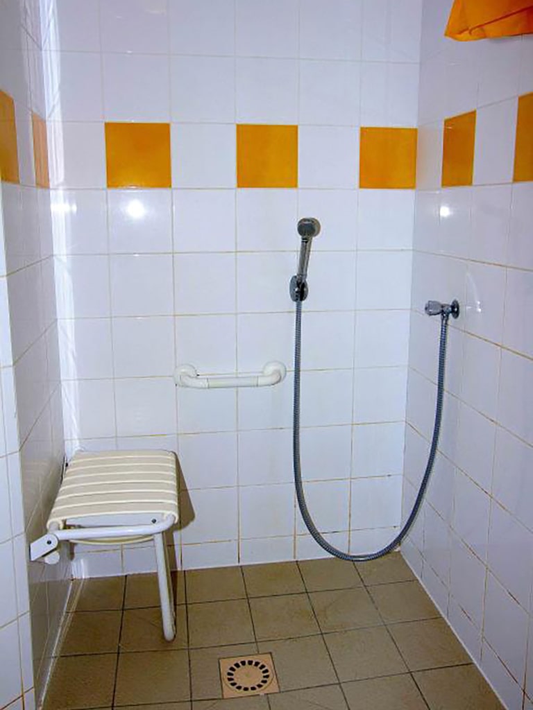 photo d'une sale de bain, carreaux blanc avec frise de carreaux jaune, douche adaptée aux personnes à mobilité réduite avec banc et poignée, du gîte de groupe et maison d'hôtes du Bas Mena