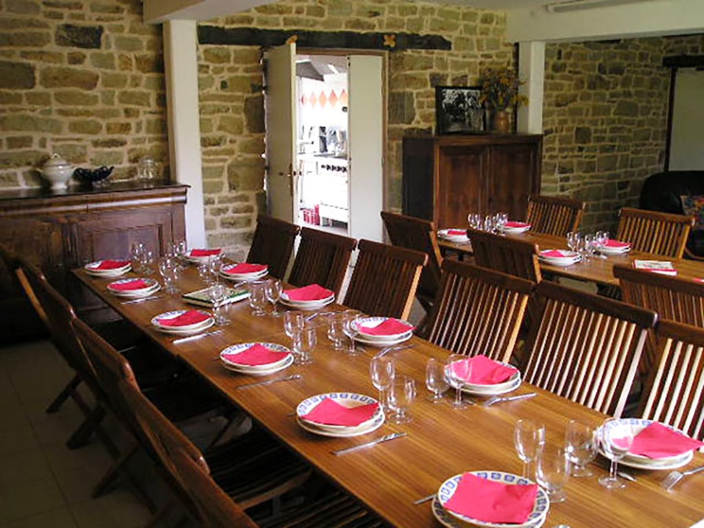 photo d'une salle à manger avec deux grandes tables et leurs chaises, deux meubles en bois foncés, mur de pierre, vue sur cuisine professionnelle dans l'entrebâillement d'une porte, du gîte de groupe et maison d'hôtes du Bas Mena