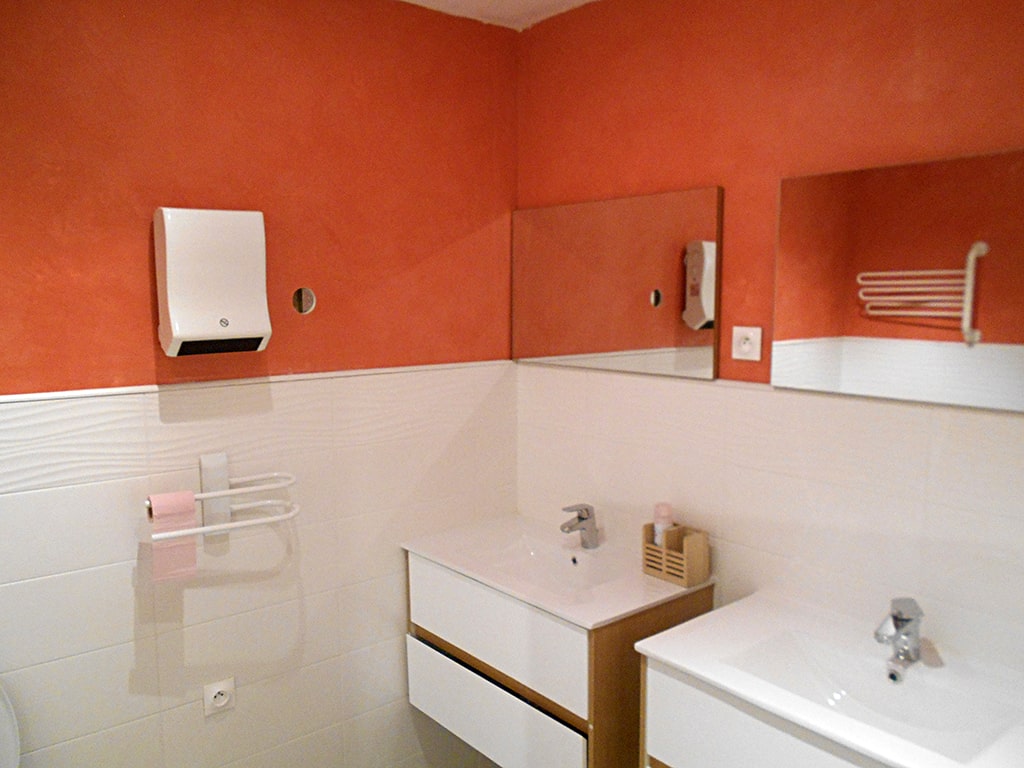 photo d'une salle de bain aux murs orange et blanc avec deux lavabos sur meubles à tiroirs, miroirs suspendus au dessus, robinet mitigeur, sèche mains et portes serviettes, prises électrique, du gîte de groupe et maison d'hôtes du Bas Mena