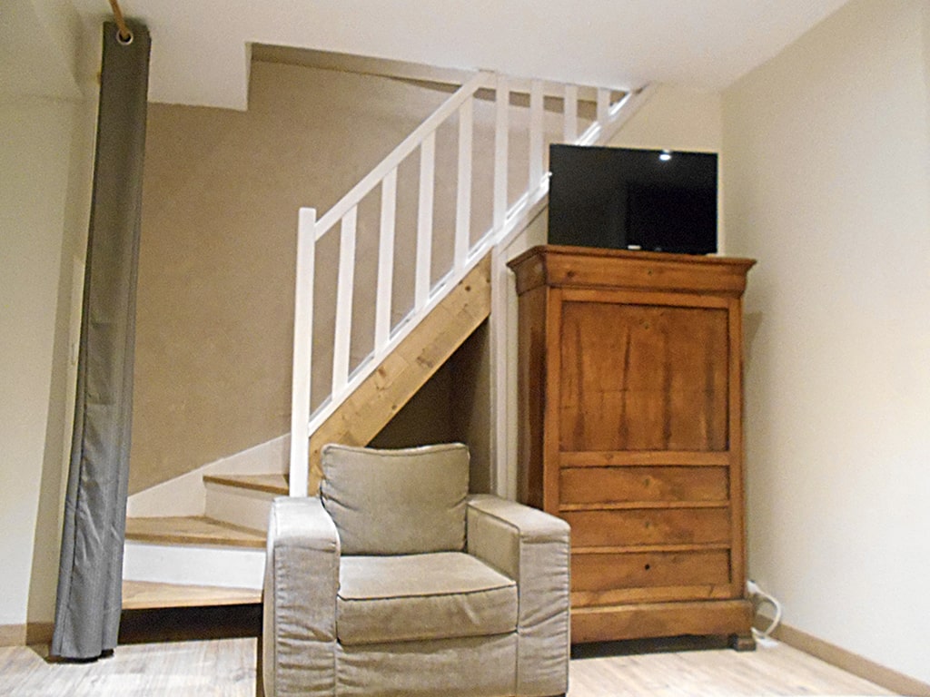 photo d'un salon avec montée d'escalier en bois derrière un fauteuil gris confortable, un haut meuble en bois avec télévision au dessus, du gîte de groupe et maison d'hôtes du Bas Mena